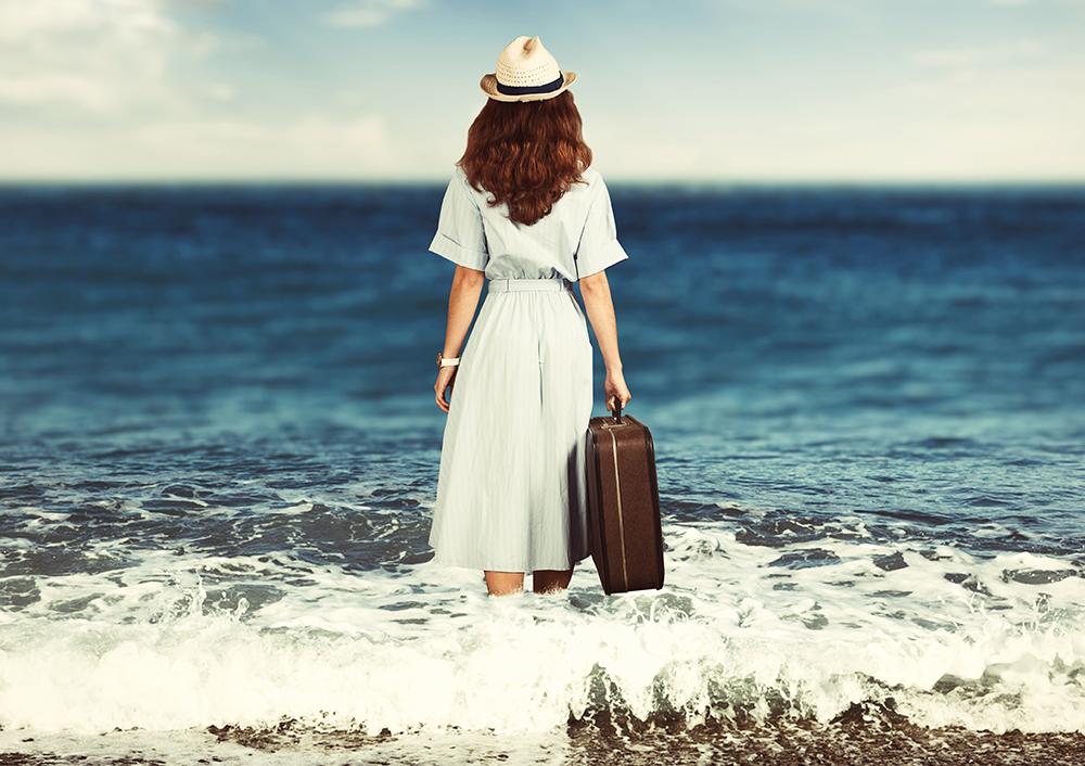 Traveler in a sun hat and light dress carrying a suitcase beside the sea